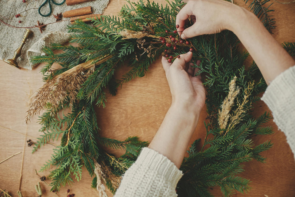 Hands holding fir branches, and pine cones, thread, berries, scissors on wooden table.