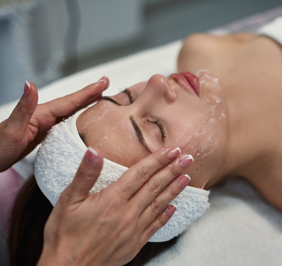 A spa worker gently applies a facial product to a woman's face with their hands. The woman is relaxed with her eyes closed.