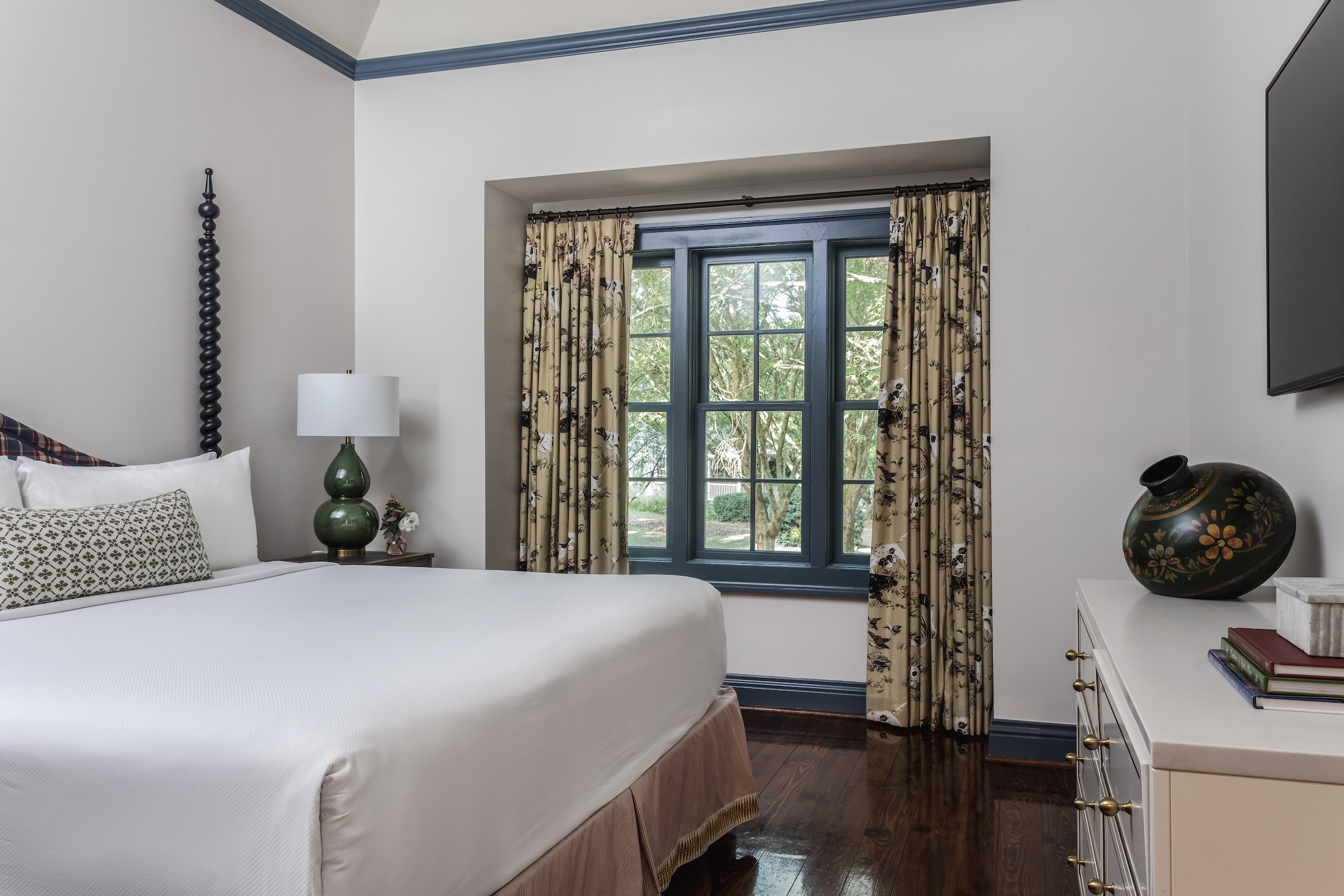 A bedroom within an Arbor Cottage at Barnsley Resort, a queen bed, wall-mounted TV, dresser, side table with lamp and a large window are pictured.