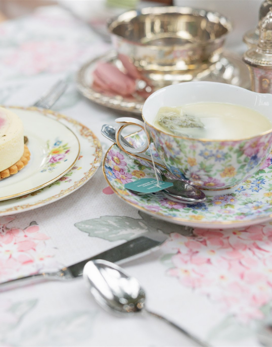 A cup of tea and plated pastry on a floral print table with flowers and silverware
