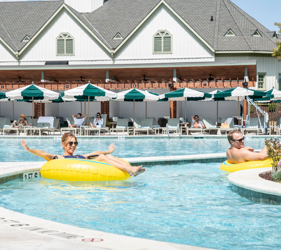 woman in a floating tube in a pool lazy river, people lounging on chairs under green and white umbrellas