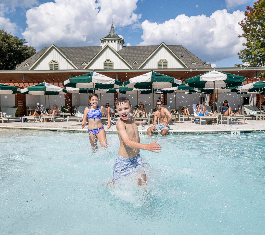 boy and girl running in shallow pool water, people lounging on chairs under green and white umbrellas