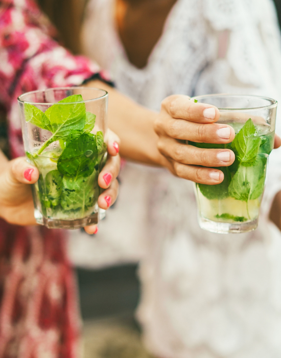 women holding out glasses on liquid with herbs inside