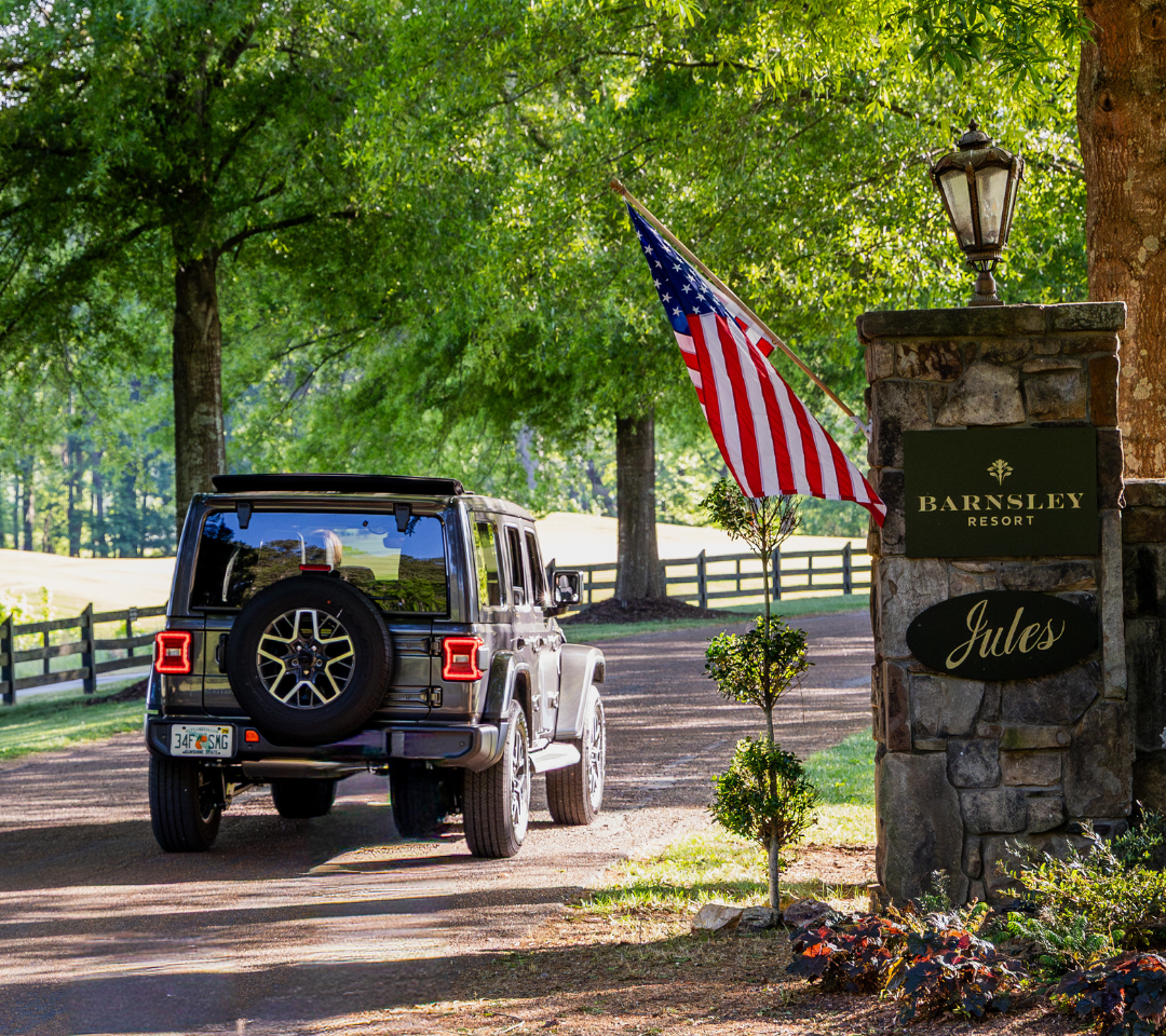 a jeep driving through an entrance of a resort on a paved fenced-lined path with an American flag hanging from stone pillar