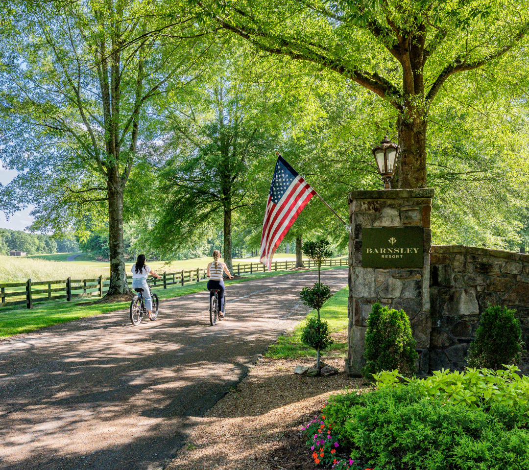 two women on bikes riding on a road through an entrance of two stone pillars, flag flying above landscaped plants