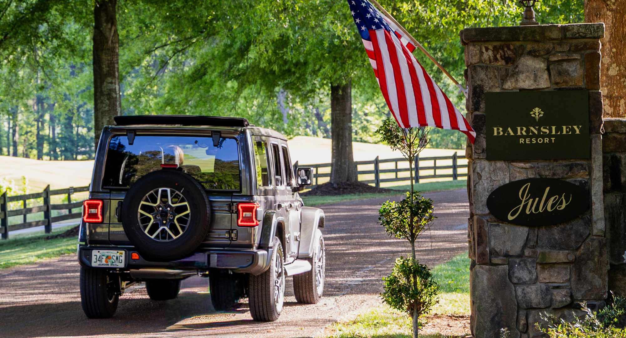 a jeep driving through an entrance of a resort on a paved fenced-lined path with an American flag hanging from stone pillar