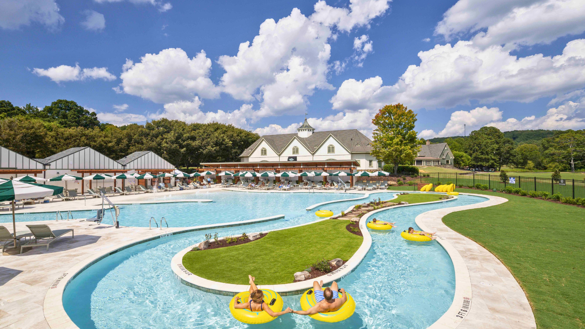 a pool with a lazy river. man and woman floating on innertubes. white building in background with cabanas and umbrellas.