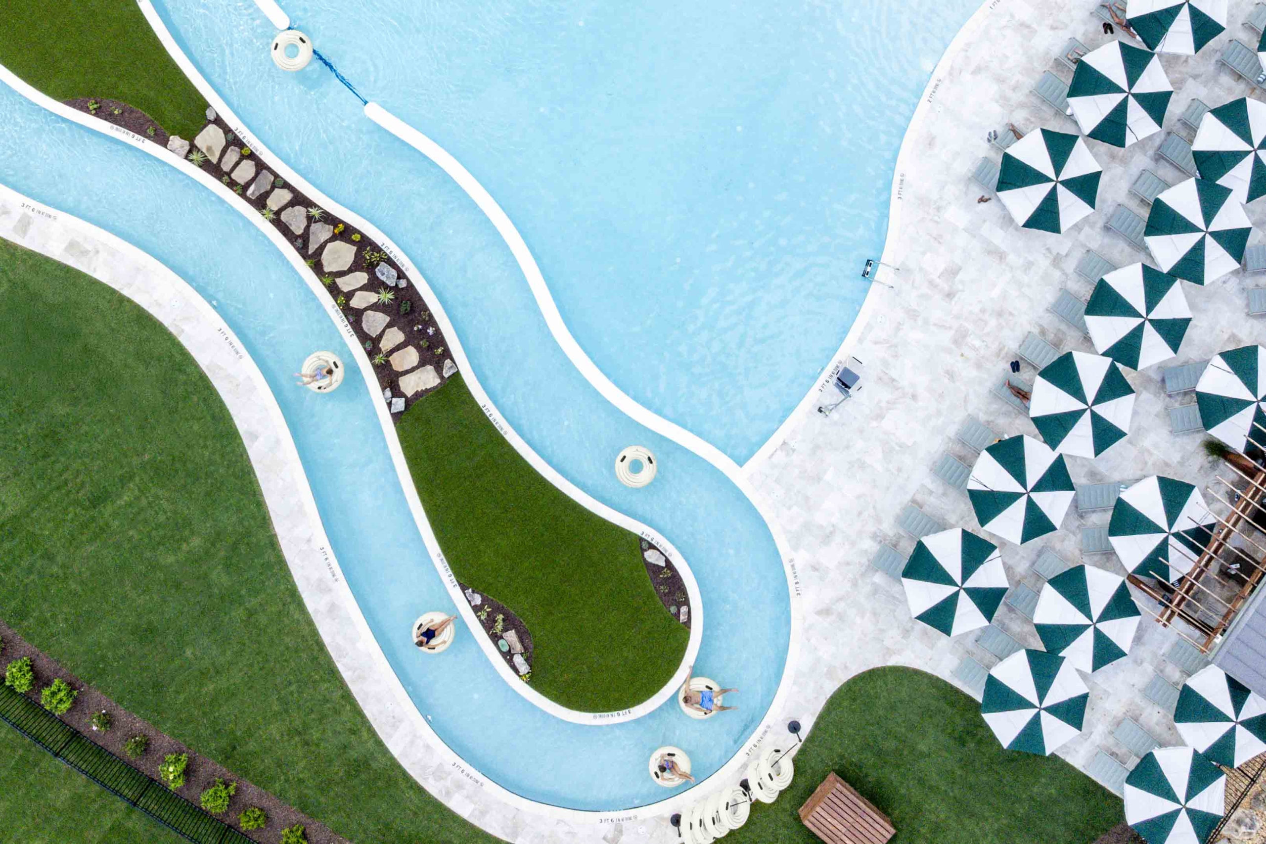 a pool with a lazy river. people floating on innertubes. white building in background with cabanas and umbrellas.