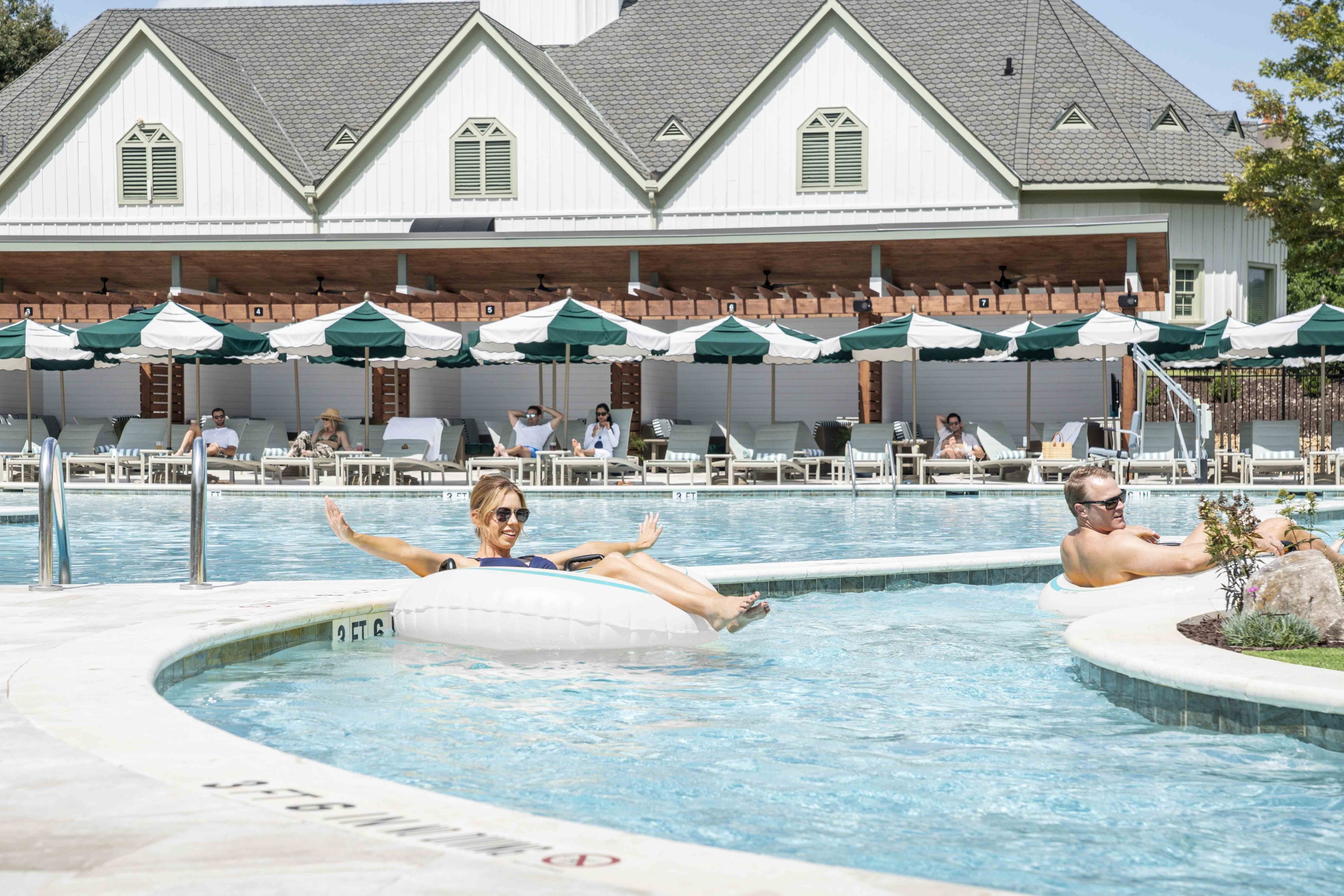 a pool with a lazy river. people floating on innertubes. white building in background with cabanas and umbrellas.