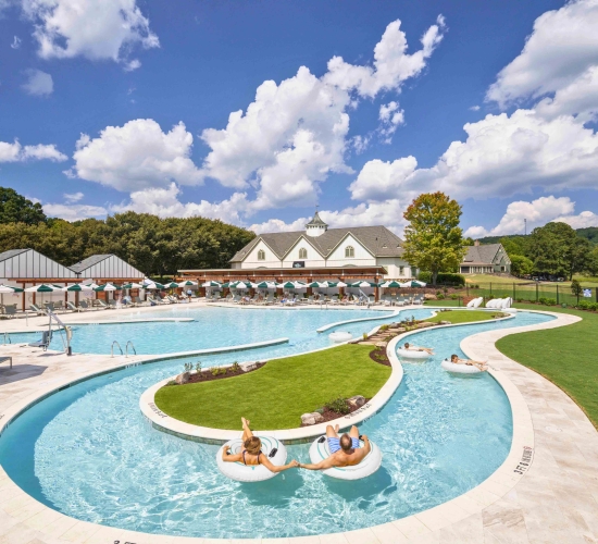 a pool with a lazy river. man and woman floating on innertubes. white building in background with cabanas and umbrellas.