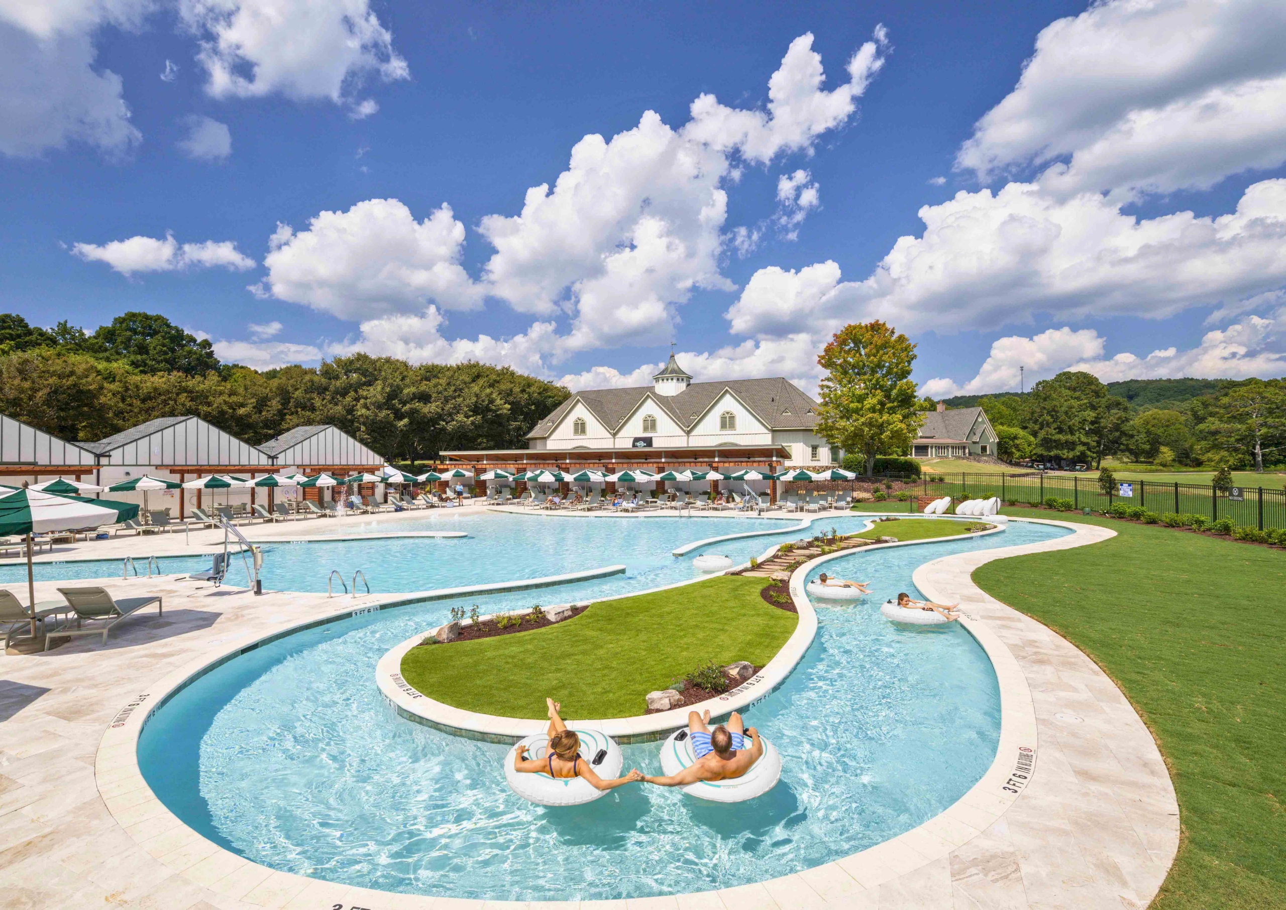 a pool with a lazy river. man and woman floating on innertubes. white building in background with cabanas and umbrellas.