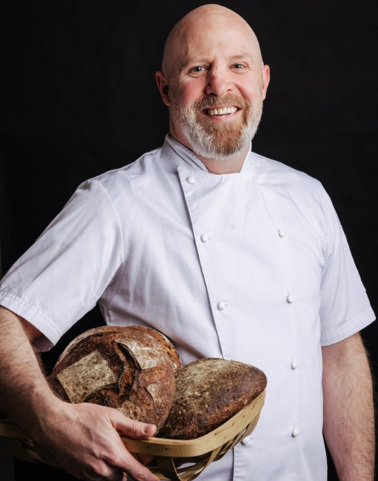man in chef's coat holding a basket of bread smiling at camera