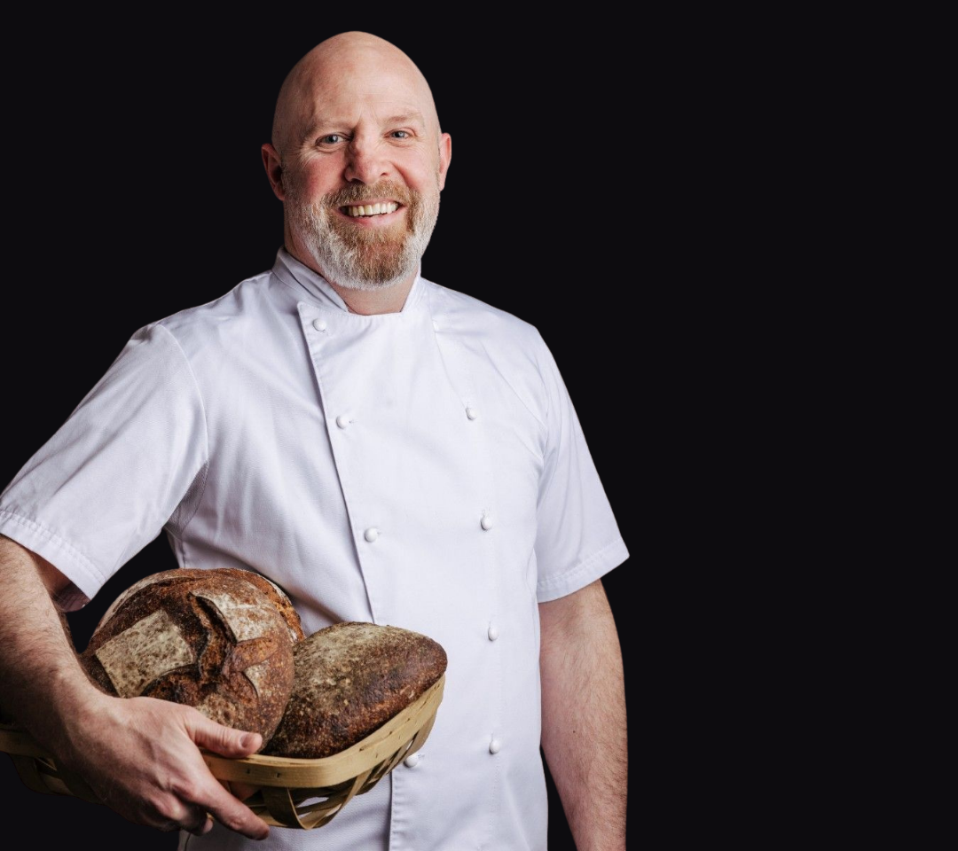 man in chef's coat holding a basket of bread smiling at camera