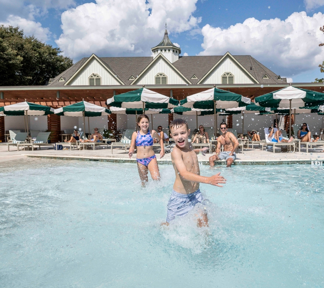 kids running through shallow water in a pool. people sitting in lounge chairs under green and white striped umbrellas.