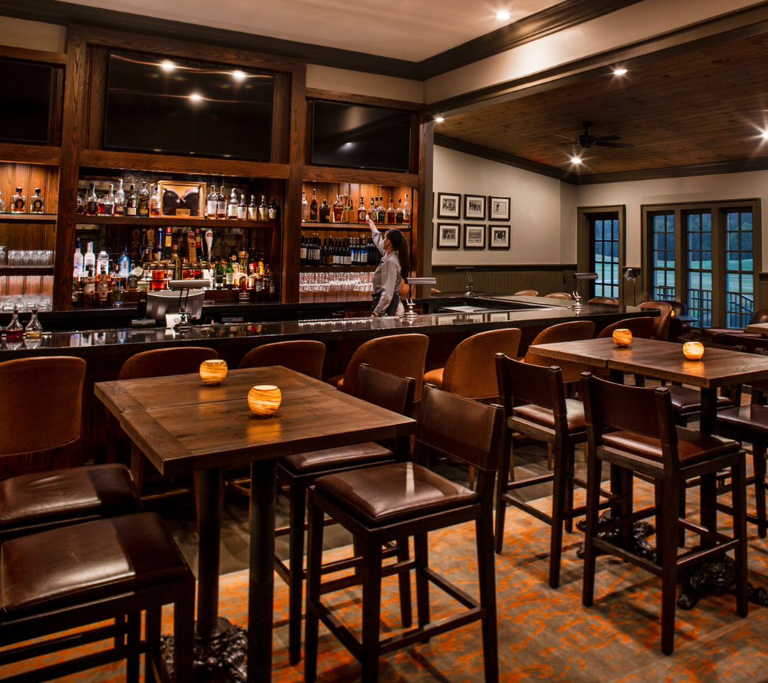 a dimly-lit bar with tables and chairs. a bartender placing a glass on a shelf