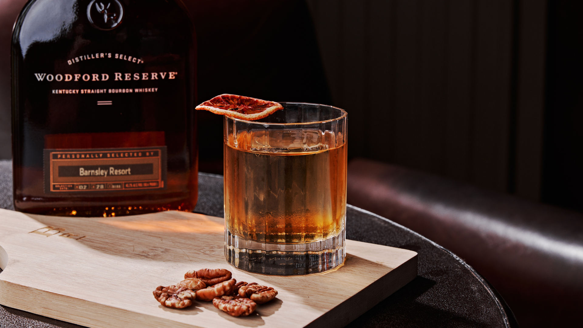 a cocktail on a wooden board with a bottle of bourbon. Pecans and sliced fruit garnish the board and glass.