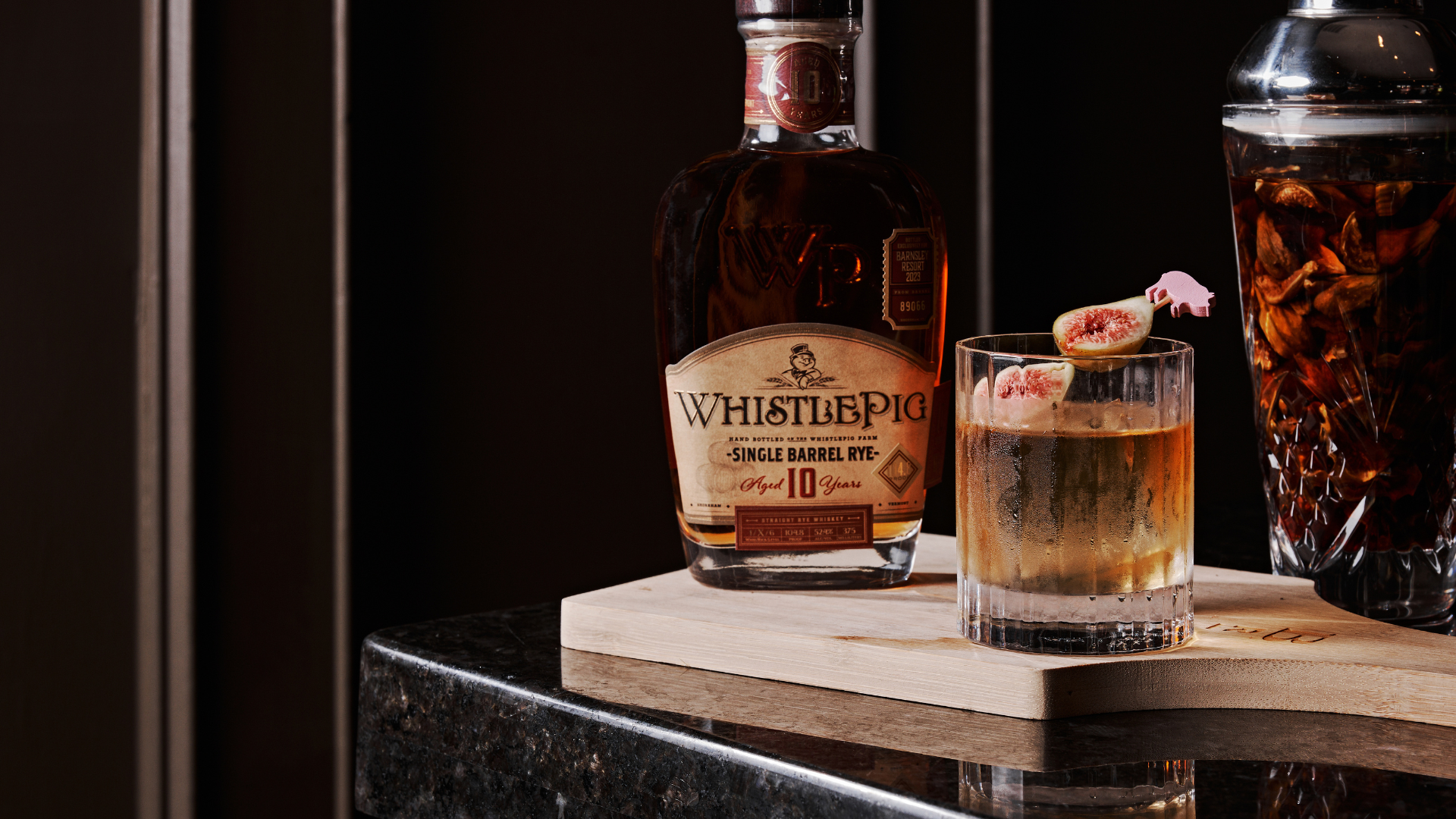 a cocktail on a wooden board with a bottle of bourbon. Fruit garnish the board and glass.