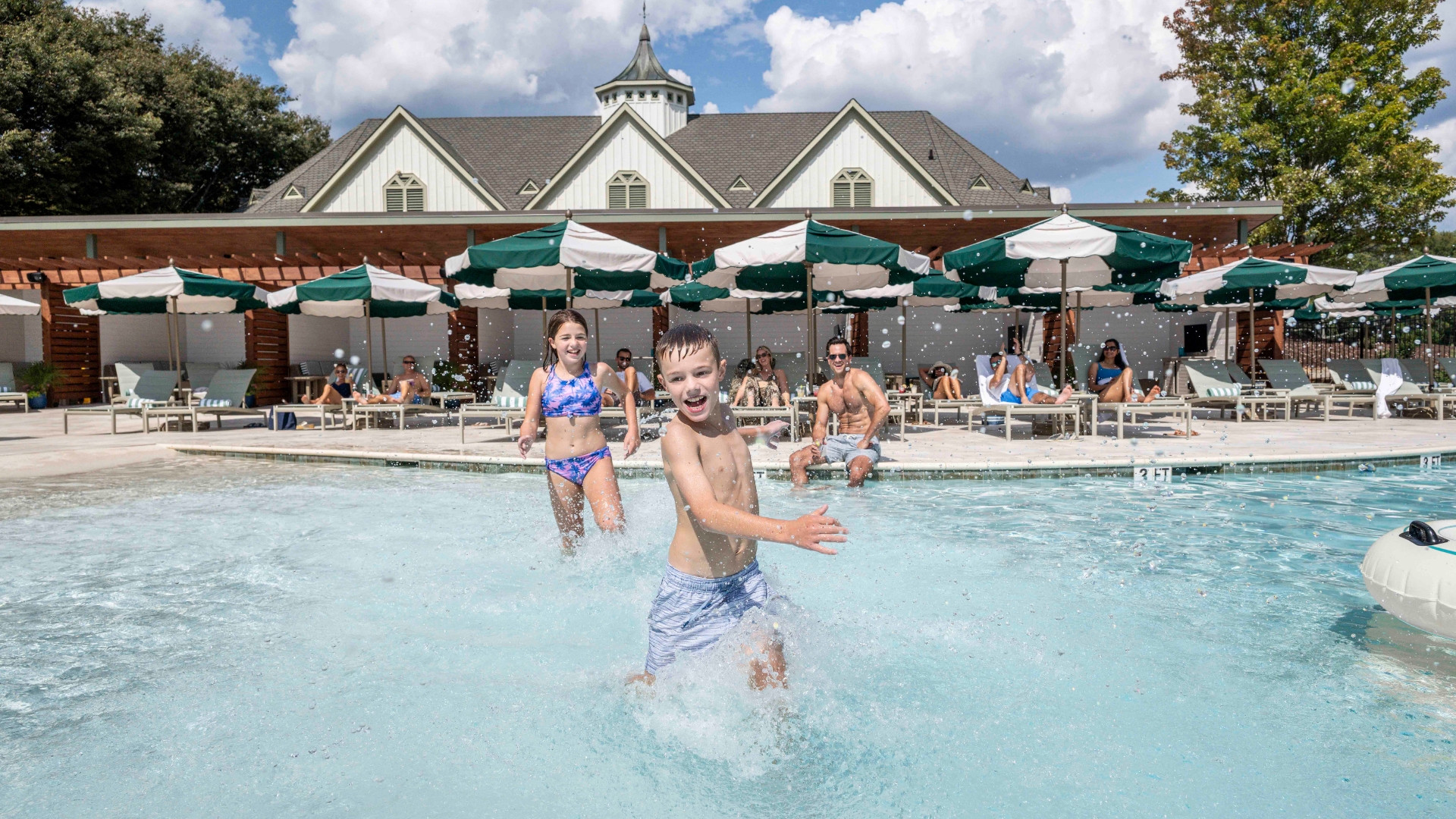 kids running through shallow water in a pool. people sitting in lounge chairs under green and white striped umbrellas.
