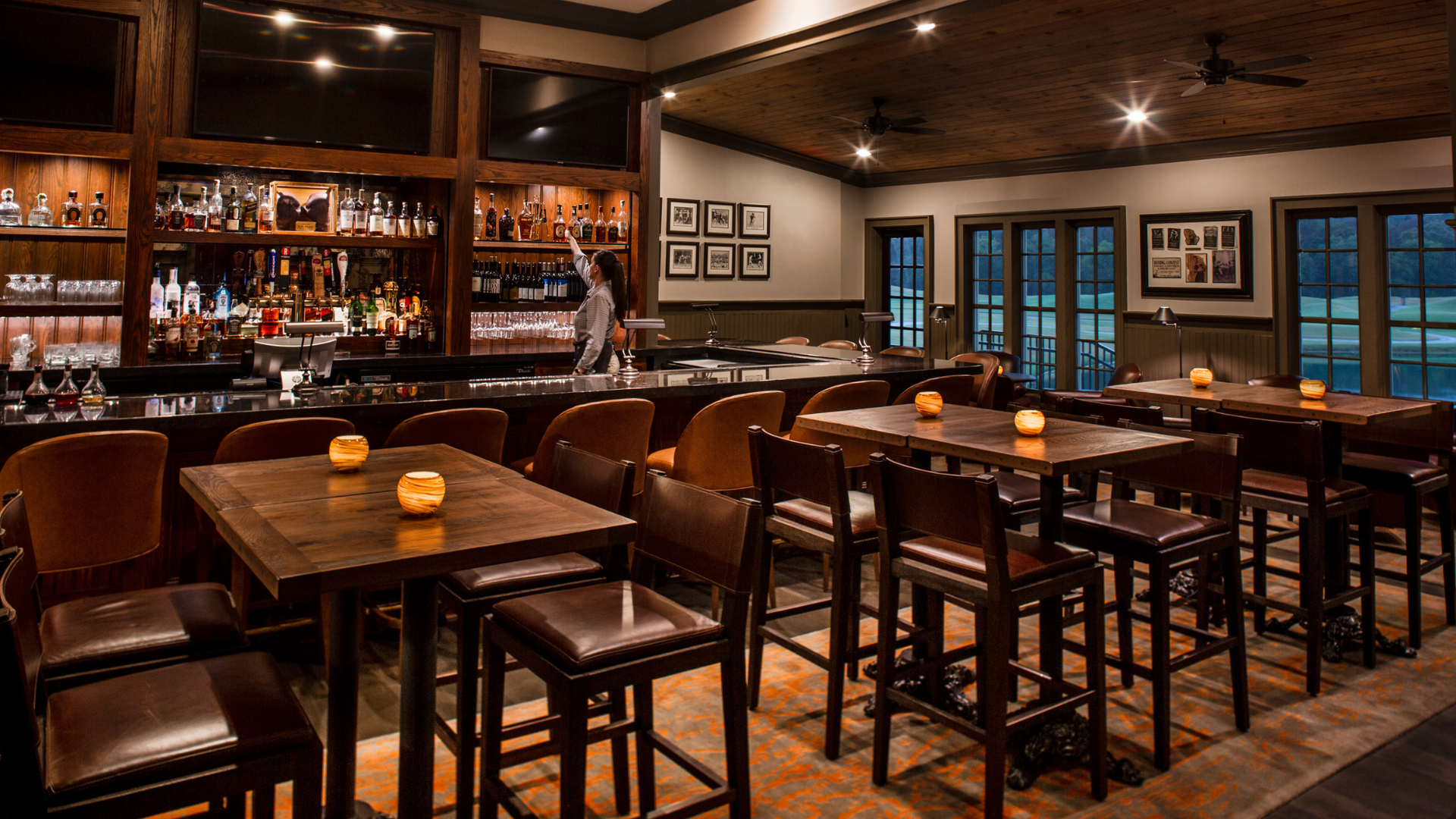 a dimly-lit bar with tables and chairs. a bartender placing a glass on a shelf