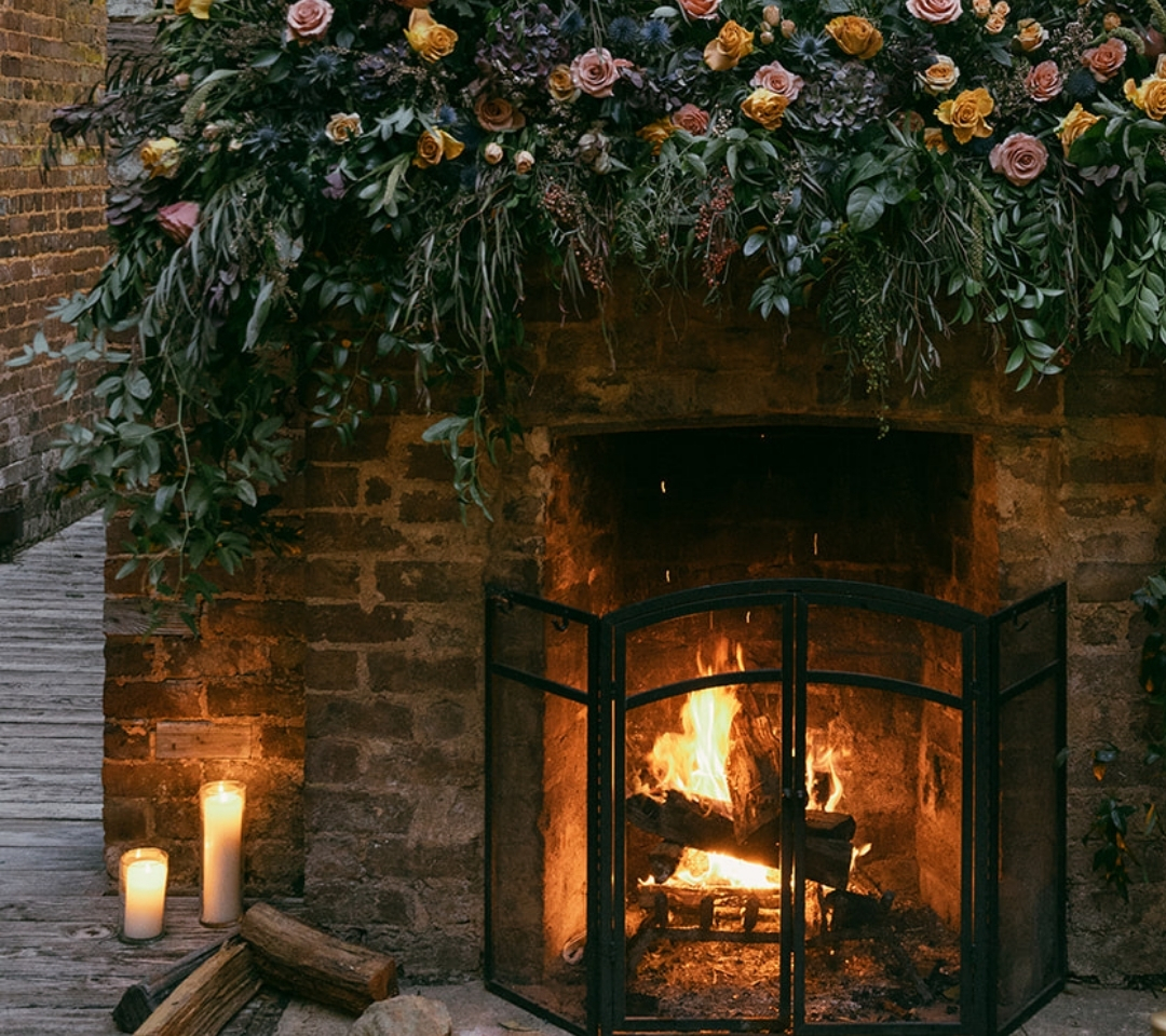 fireplace lit with fire and the mantel has flowers and greenery on top. candles sit on the floor