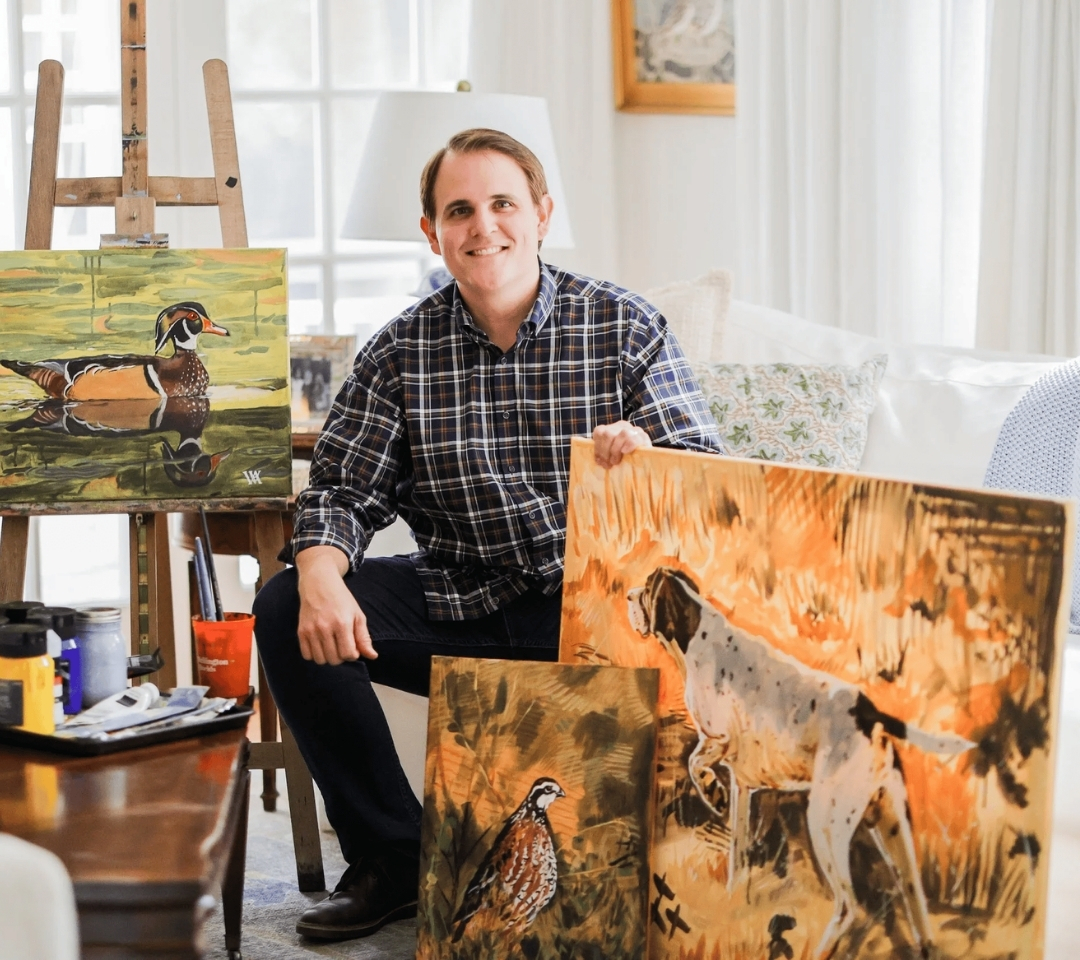 man smiling at camera, surrounded by paintings and art supplies