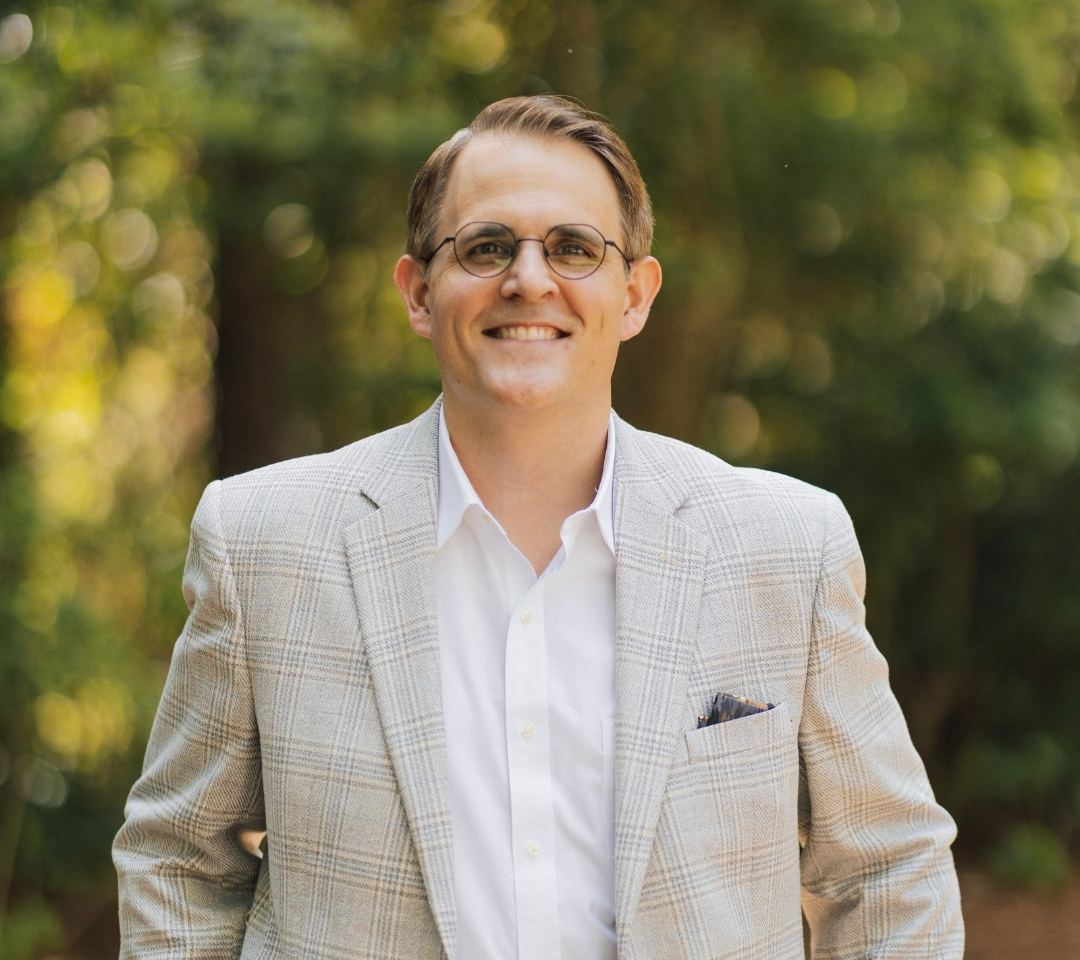 a man smiling, wearing a tan suit and glasses